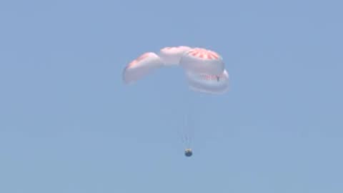 Capsule descending under parachutes in clear blue sky.