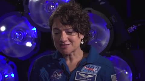 Speaker in blue flight suit talking in front of illuminated blue backdrop.