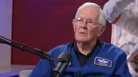An elderly man in glasses and a blue NASA jacket speaking in an interview