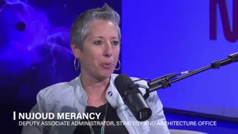 A woman with short grey hair speaks during an interview against a blue background.