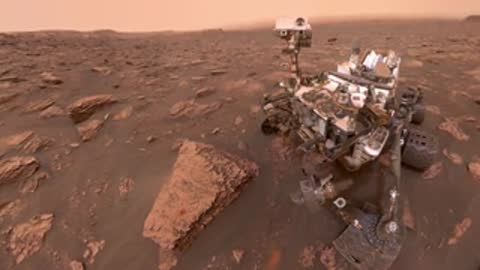 A Mars rover sits on the reddish-brown rocky surface of the Martian landscape.