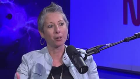 A woman with short grey hair speaks with hand gestures during an interview.
