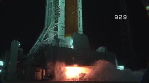 Flames ignite at the base of a large space rocket during a night launch.