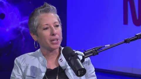 A woman with short grey hair speaks during an interview in front of a blue backdrop.
