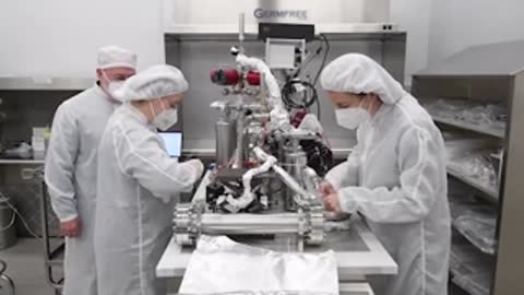 Workers in white cleanroom suits assemble aerospace equipment at a workbench.