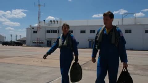 Two astronauts in blue jumpsuits walk across the tarmac near a building.
