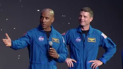 Two astronauts in blue jumpsuits stand on stage in front of a starry background.