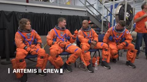 Four astronauts in orange spacesuits sit in a row and talk.