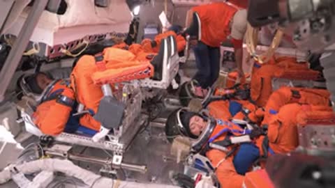 Astronauts in orange spacesuits lie inside a spacecraft simulator being assisted.
