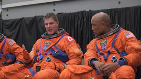 Two astronauts in orange spacesuits sit and converse during a briefing.