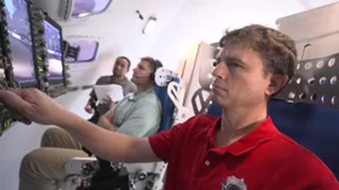 A man in a red shirt gestures with his hand inside a flight simulator.