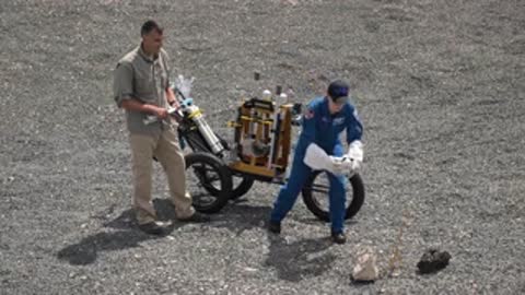 An astronaut and a support worker operate a cart with gas cylinders outdoors.