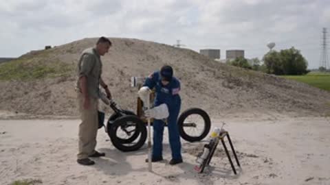 An astronaut and support staff work with equipment near a dirt mound.