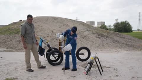 An astronaut and worker stand near equipment and a bicycle on dirt.