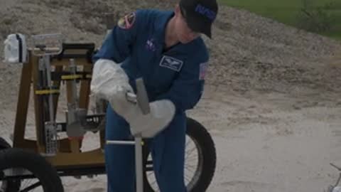 An astronaut adjusts mechanical equipment outdoors near a dirt mound.