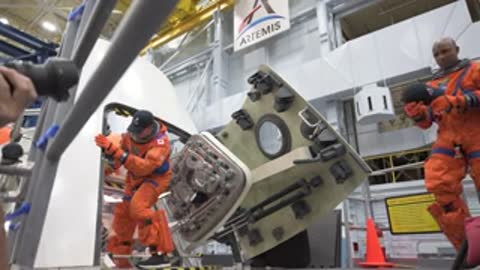 Astronauts in orange spacesuits exit a spacecraft mockup in a facility.