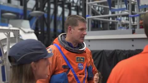 Astronauts in orange spacesuits converse with support staff outdoors.