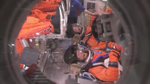 An astronaut in an orange spacesuit rests inside a spacecraft cabin simulator.