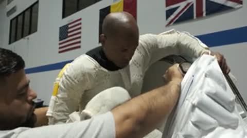 Astronaut puts on spacesuit with assistance from technicians in indoor facility.