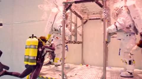 Divers assist astronaut in spacesuit inside underwater training facility pool.