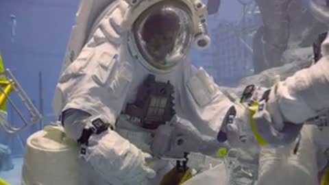 Two astronauts in spacesuits interact and adjust equipment during underwater training session.