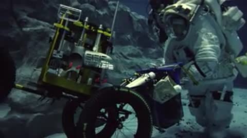 Astronaut pushes wheeled equipment cart across underwater floor during simulation.