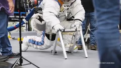 Astronaut kneels and adjusts foldable metal frame structure on indoor floor.