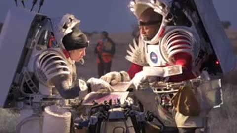 Two astronauts in spacesuits examine documents and equipment outdoors at dusk.