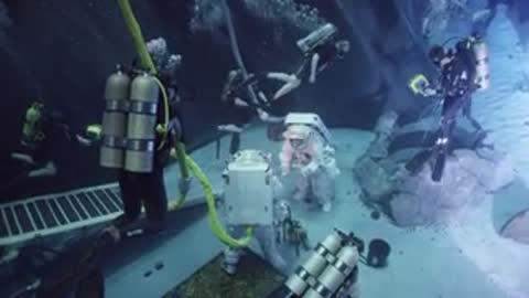 Divers and astronaut operate underwater near rocky structure during training simulation.