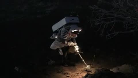 Astronaut uses metal detector and flashlight outdoors at night near vegetation.