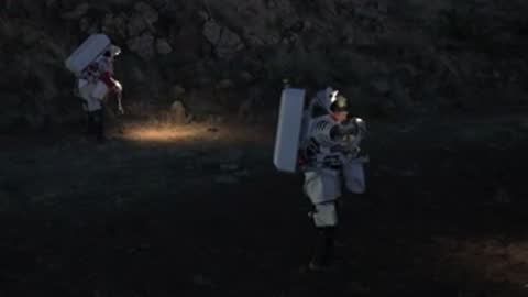 Astronauts in space suits walk across a dark landscape lit by portable lights.