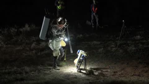 An astronaut in a space suit works near equipment outdoors at night.