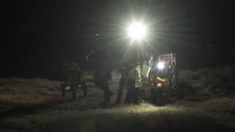 Astronauts stand behind equipment emitting a bright glare in a dark setting.