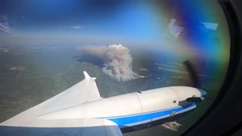 Aerial view through airplane window showing smoke from a fire below.