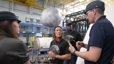 Instructor explaining camera equipment to astronauts beneath a hanging moon model.