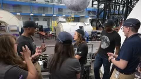 Astronauts and instructors talking and gesturing around a hanging moon model.