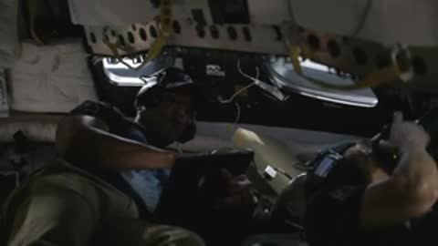 Astronaut reaching across seats inside a spacecraft simulator cabin.