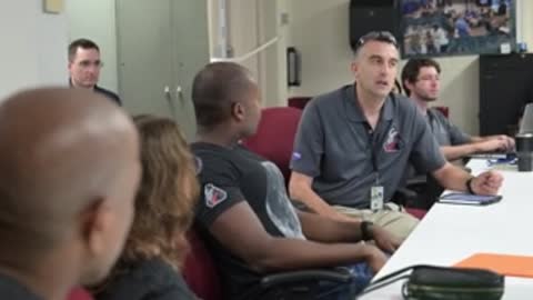 Astronauts listening as a man in a grey polo speaks at a table.