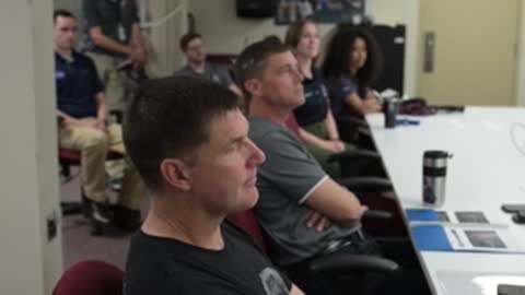 An astronaut sits in profile view inside a conference room.