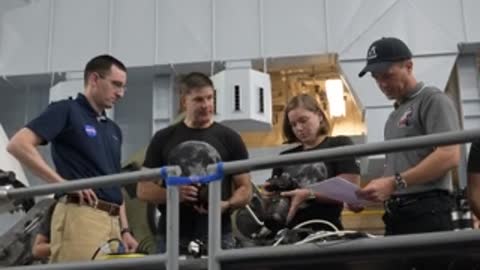 Astronauts examine documents near a simulator setup in a hangar.