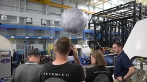 Astronauts stand near a hanging moon model inside a facility.