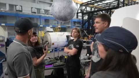 Astronauts talk near a spacecraft simulator in a large room.
