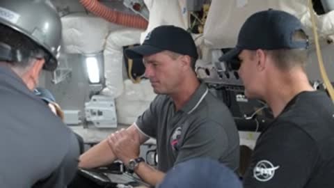 Two astronauts sit inside a spacecraft simulator cockpit talking.