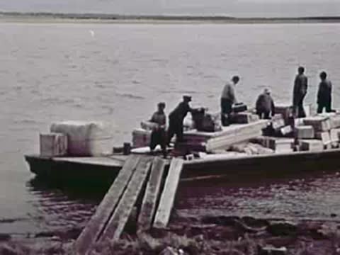 Workers loading cargo onto a barge from a wooden ramp.