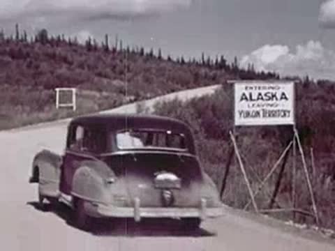 A car drives past a sign that reads "Entering Alaska, Leaving Yukon Territory".