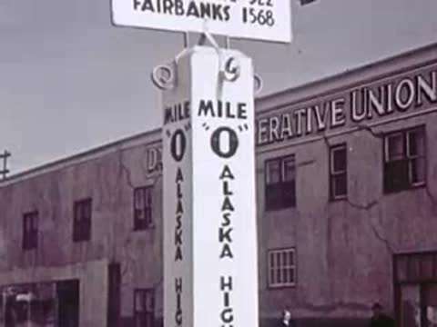 A close-up of a Mile 0 signpost for the Alaska Highway in front of a building.