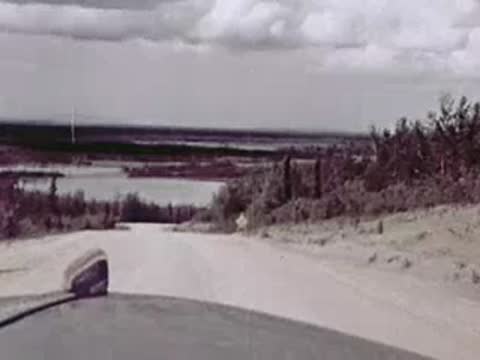 Looking ahead from a moving vehicle down a sloping gravel road toward water and trees.