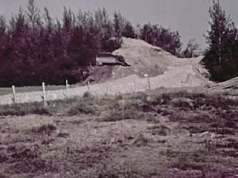 A dirt mound with a metal drainage pipe sits beside a rural road surrounded by trees.