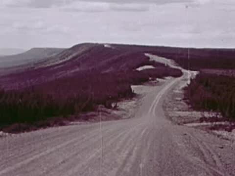 A vehicle drives along a gravel road cutting through a rolling, forested landscape.