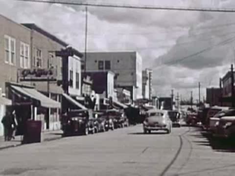 A town street lined with parked vintage cars, storefronts, and overhead utility lines.